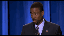 a man in a suit and tie with an american flag on his chest stands in front of a blue curtain