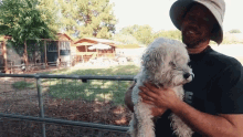 a man in a hat holds a small white dog in his arms with the letter e on his shirt