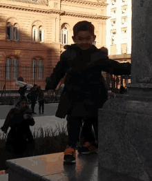 a little boy wearing a black jacket and orange shoes is standing on a ledge in front of a building