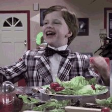 a young boy in a plaid suit and bow tie is sitting at a table with a plate of food .