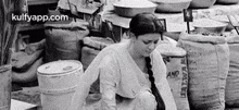 a black and white photo of a woman sitting in front of a pile of bags in a market .