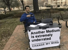 a man is sitting at a table with a sign that says `` another medium is extremely underrated change my mind '' .