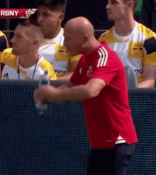 a man in a red shirt is holding a bottle of water in front of a screen that says rbny