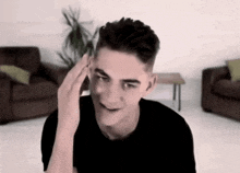 a young man in a black shirt is smiling in a living room while touching his head .