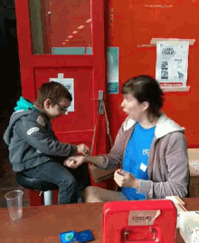 a woman sits at a table talking to a young boy who is wearing a blue shirt that says ' boys '