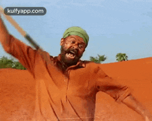 a man with a beard and a green turban is holding a stick in front of a red sand dune .