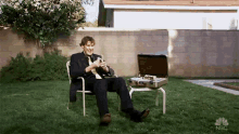 a man in a suit and tie is sitting in a chair in a backyard with a briefcase on a table .