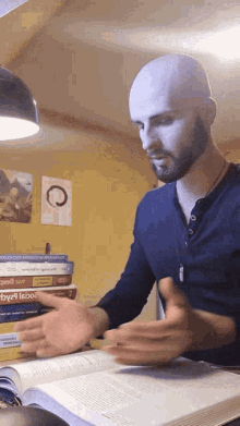 a bald man with a beard is sitting at a table reading a book with a stack of books on it