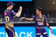 two cricket players are standing next to each other on a field . one of the players is wearing a purple jersey .