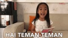 a little girl is sitting on a couch with hai teman-teman written on the bottom