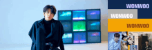 a man in a black coat is standing in front of a wall of televisions with the name wonwoo written on it