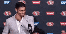 a man in a suit is standing in front of a microphone with a san francisco 49ers logo on the wall behind him .