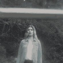 a woman in a denim jacket is standing in front of trees