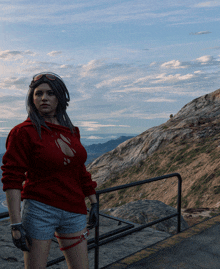 a woman in a red sweater and shorts stands on a ledge overlooking a mountain