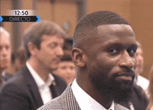 a man in a suit and tie is sitting in front of a screen that says 12:50 directo