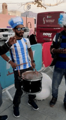 a man in a blue and white striped shirt playing a drum in front of a now bus