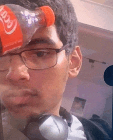 a man with glasses and headphones is holding a bottle of coca cola on his forehead .