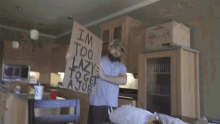 a man in a kitchen holds up a cardboard sign that says i 'm too lazy to get a job