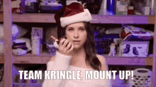 a woman wearing a santa hat is holding a candy cane and says team kringle mount up .