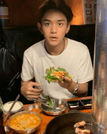 a young man is sitting at a table eating food with a bottle of wine behind him