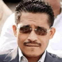 a man with a mustache wearing sunglasses and a suit is looking at the camera .