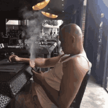 a bald man is sitting at a table smoking a cigar while looking at his phone .