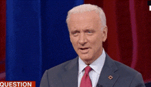 a man in a suit and tie is standing in front of a red curtain and a sign that says question .