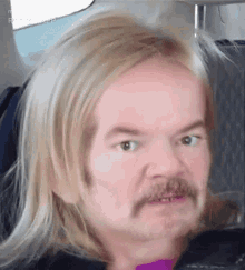 a man with long blonde hair and a mustache is sitting in a car .