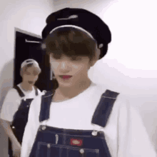 a boy wearing overalls and a beret is standing next to another boy wearing overalls .