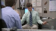 a man in a green shirt and blue tie is sitting at a desk in front of a computer monitor .