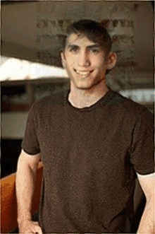 a young man wearing a brown t-shirt is standing in front of a window and smiling .