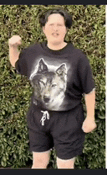 a woman wearing a wolf t-shirt and shorts is standing in front of a bush .