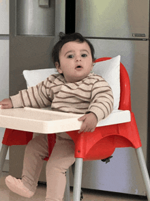 a baby is sitting in a red high chair in front of a stainless steel refrigerator