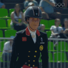 a woman wearing a helmet stands in front of a crowd and the olympic rings are visible in the background