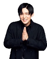 a young man in a black suit is smiling with his hands folded in prayer .