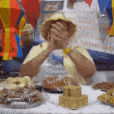 a woman in a yellow hat is covering her face with her hands while sitting at a table with plates of food .