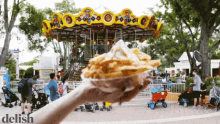 a person is holding a container of french fries in front of a carousel and the word delish is on the bottom right