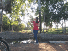 a woman in a red shirt and blue jeans stands in front of trees