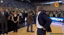 a man in a suit stands on a basketball court in front of a crowd and a sign that says tion