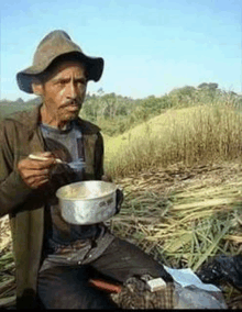 a man in a cowboy hat is sitting in a field eating from a pot