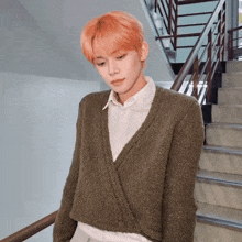 a young man with orange hair is standing next to a set of stairs wearing a green sweater .