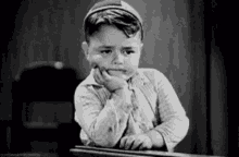 a black and white photo of a young boy sitting at a desk with his hand on his face .