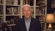 a man in a suit and plaid shirt stands in front of a bookshelf that has a picture of a man on it