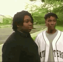 two young men are standing next to each other on a sidewalk . one of the men is wearing a baseball jersey .