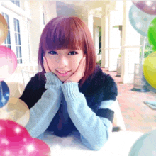 a woman in a blue sweater sits at a table with her hands on her face in front of balloons