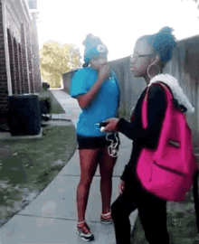 two girls are standing on a sidewalk one is wearing a blue shirt and the other is wearing a pink bag