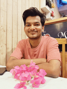 a man in a pink shirt sits at a table with flowers in front of a menu board