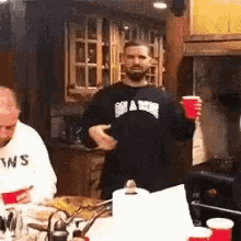 a man wearing a black shirt with the word shark on it is holding a red cup in a kitchen .