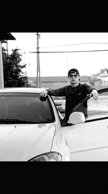 a man wearing sunglasses and a black hat is leaning against a white car