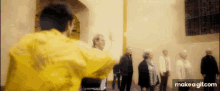 a group of people are standing in a room and a man in a yellow jacket is standing in front of them .